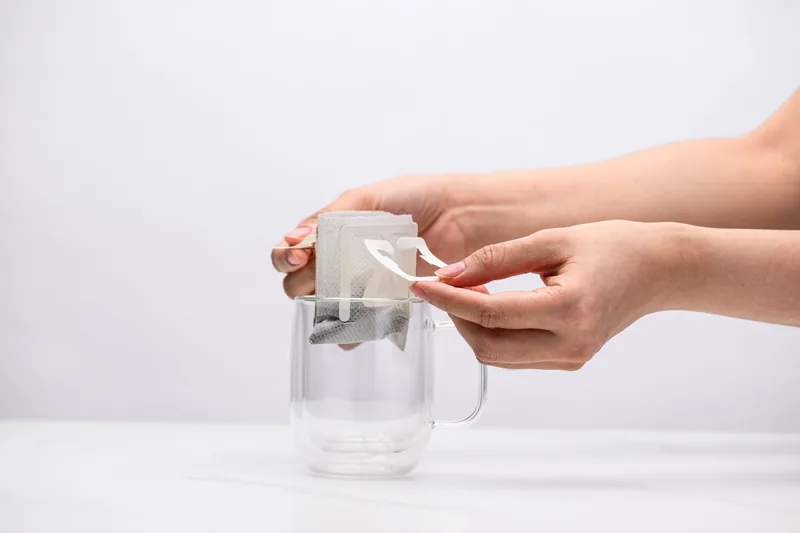 Two hands hooking the paper arms of a Sampan drip bag over the rim of a clear glass mug